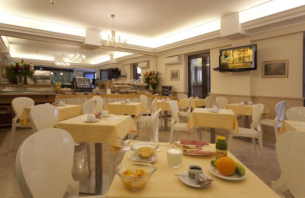 A dining area with multiple tables and chairs, a television mounted on the wall, and a kitchen in the background.