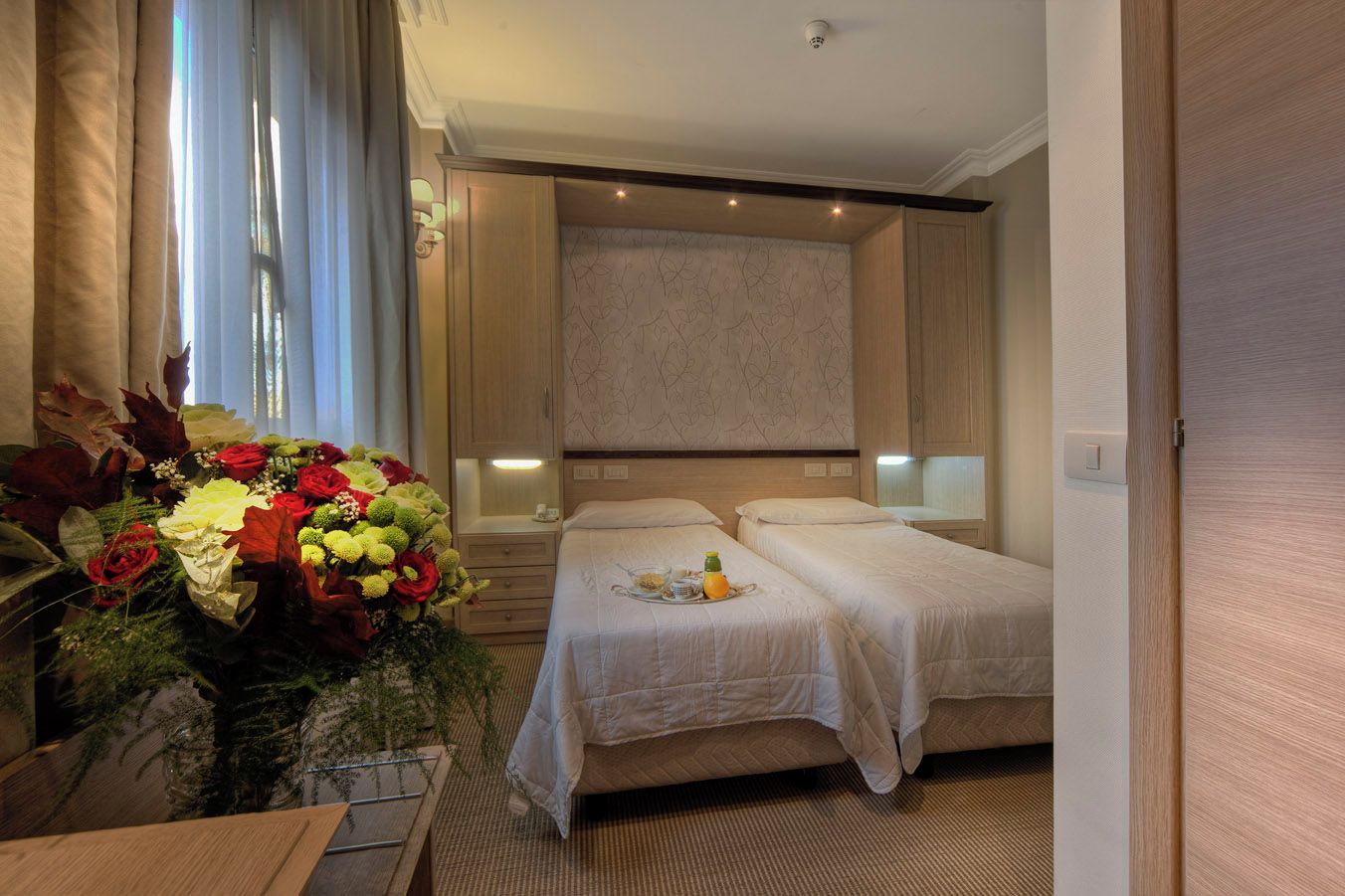 Habitación de hotel con dos camas, un ventanal con cortinas, un arreglo floral y una mesita con bandeja.