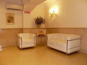 A lobby with two white armchairs, a small table with a vase of flowers, and a wall-mounted light fixture. - Galileo (Rome)