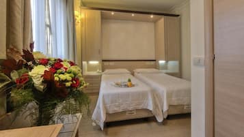 Habitación de hotel con dos camas, una mesita de noche con lámpara, un arreglo floral grande y una ventana con cortinas.