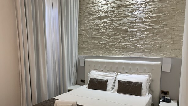 A neatly made bed with white and brown linens, a stone-textured wall behind, and a white nightstand with a phone.