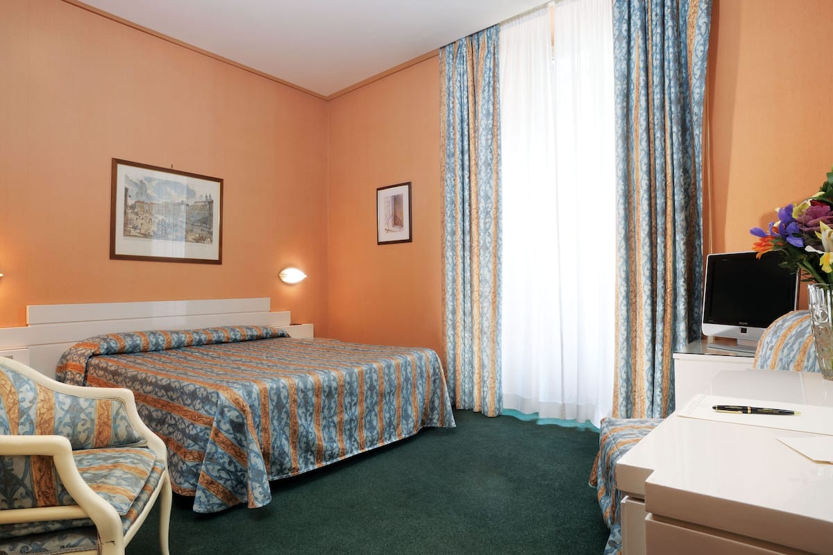 Et hotelværelse med en seng, to stole, et skrivebord, et fjernsyn og billeder i rammer på væggen.