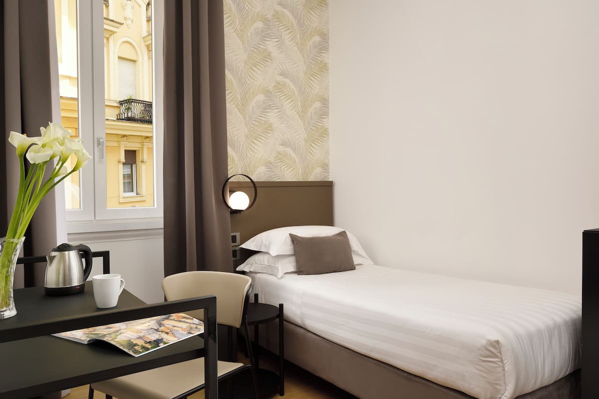 Et hotelværelse med seng, skrivebord med stol, kaffemaskine, vase med blomster og et vindue med udsigt til bygninger.