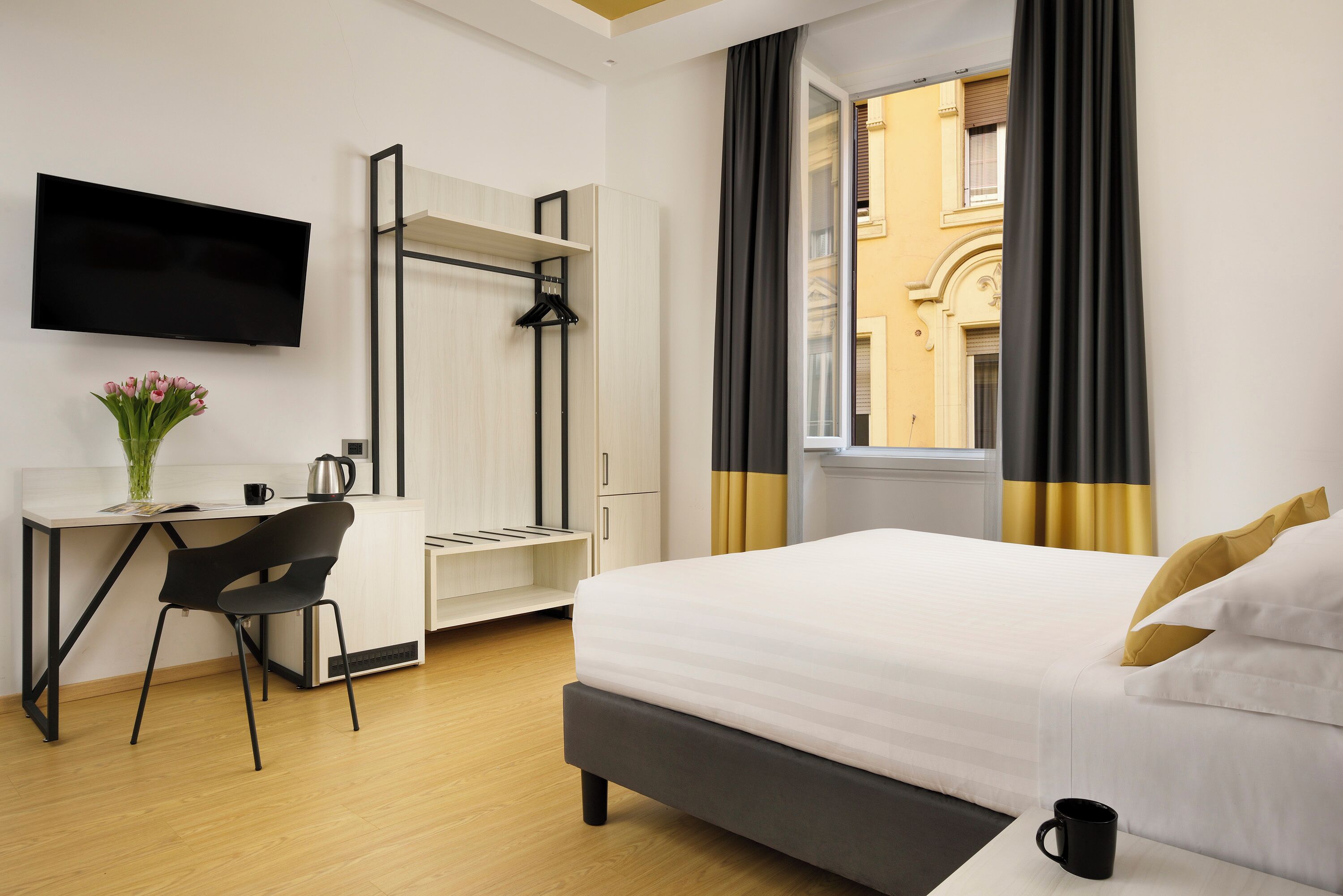 A hotel room with a bed, a desk with a chair, a TV mounted on the wall, and a window with curtains.