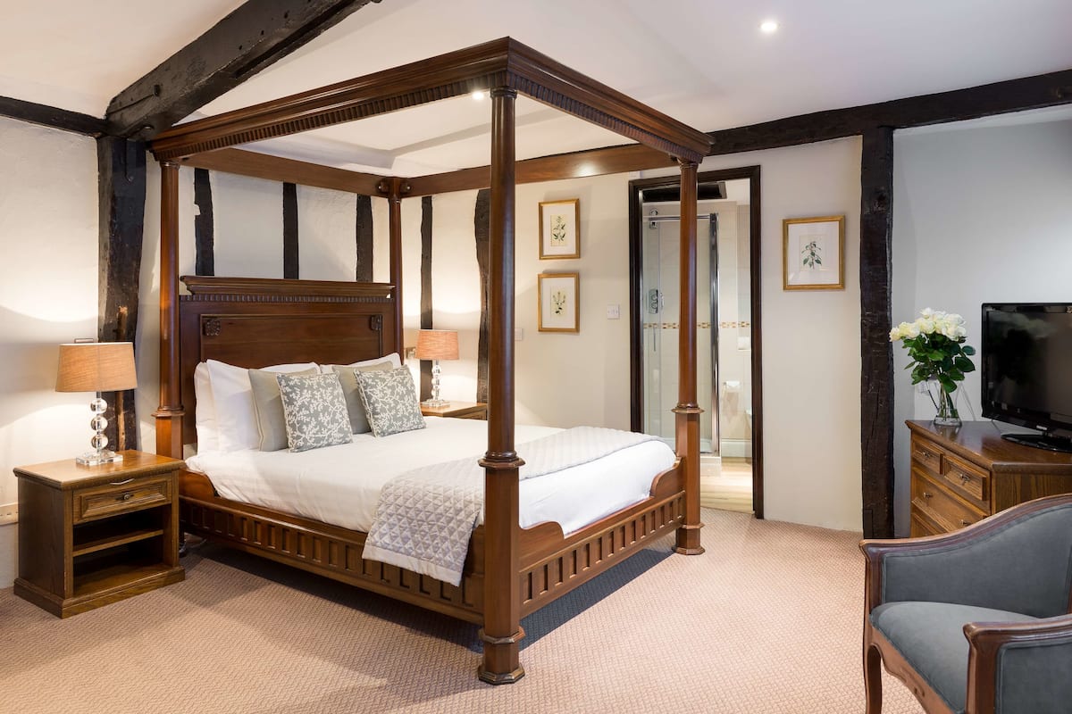 A four-poster bed with a wooden frame, a nightstand with a lamp, a television on a wooden stand, and a chair.