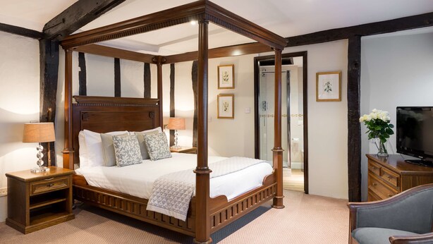 A four-poster bed with a wooden frame, a nightstand with a lamp, a television on a wooden stand, and a chair.