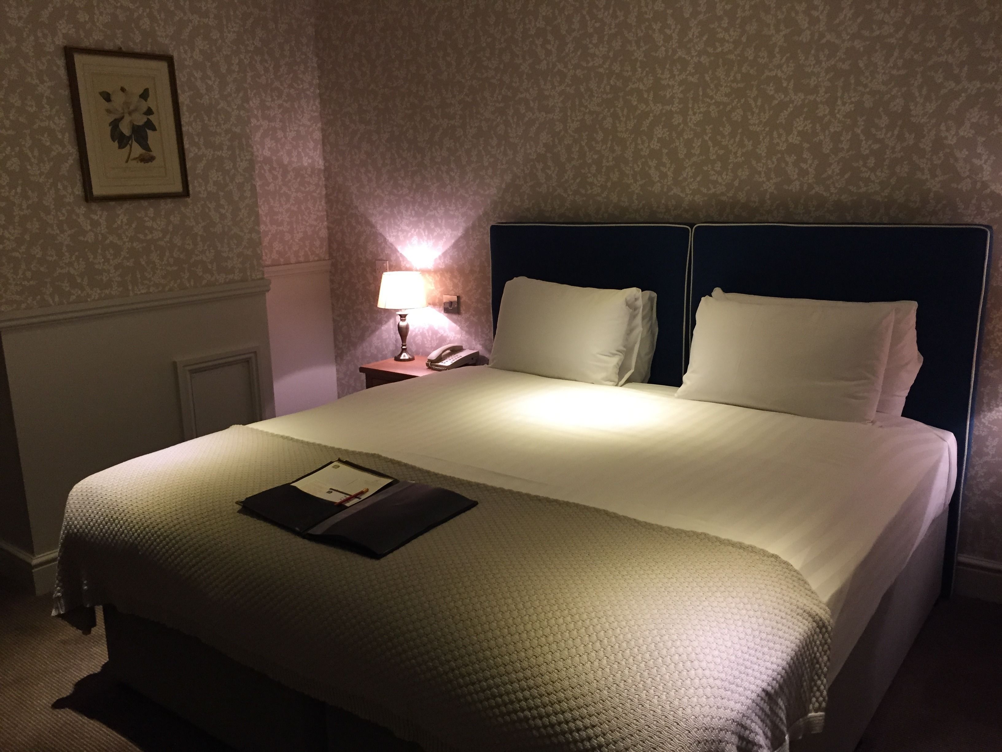Una cama bien tendida con una colcha blanca y almohadas, una lámpara de noche y un cuadro en la pared.