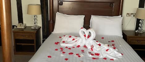 Una cama bien hecha con cabecera de madera, sábanas blancas y pétalos de rosa dispuestos en forma de cisne.