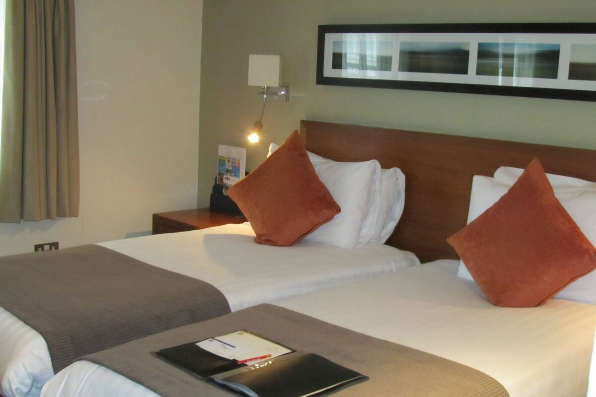 A hotel room with two beds, a nightstand with a lamp, a framed picture on the wall, and a window with curtains.