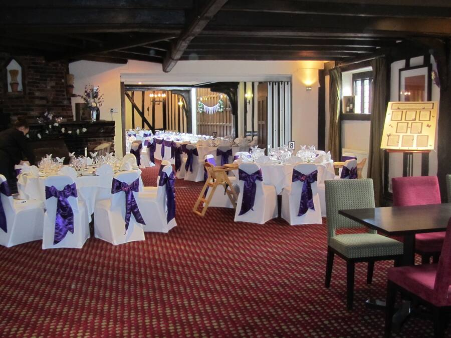 Een feestzaal met ronde tafels, bedekt met witte tafelkleden en paarse linten, in rijen opgesteld.