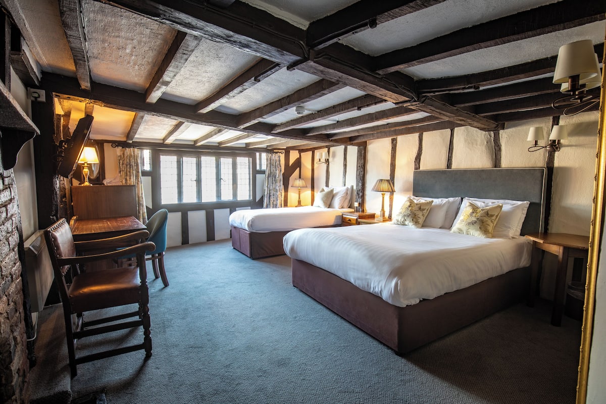 A traditional room with exposed wooden beams, two single beds, a wooden chair, and a desk with a lamp.