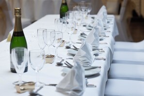 Een lange eettafel, gedekt met witte tafelkleden, drinkglazen en bestek.