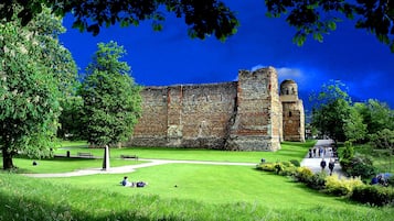 A historic stone building with a grassy courtyard, surrounded by trees and a clear blue sky.