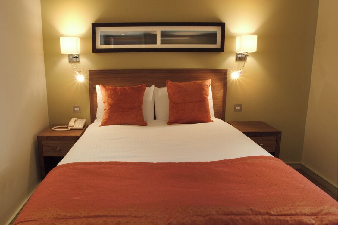Ein Hotelzimmer mit einem großen Bett, zwei Kissen, einem Nachttisch mit Lampe und einem gerahmten Bild an der Wand.