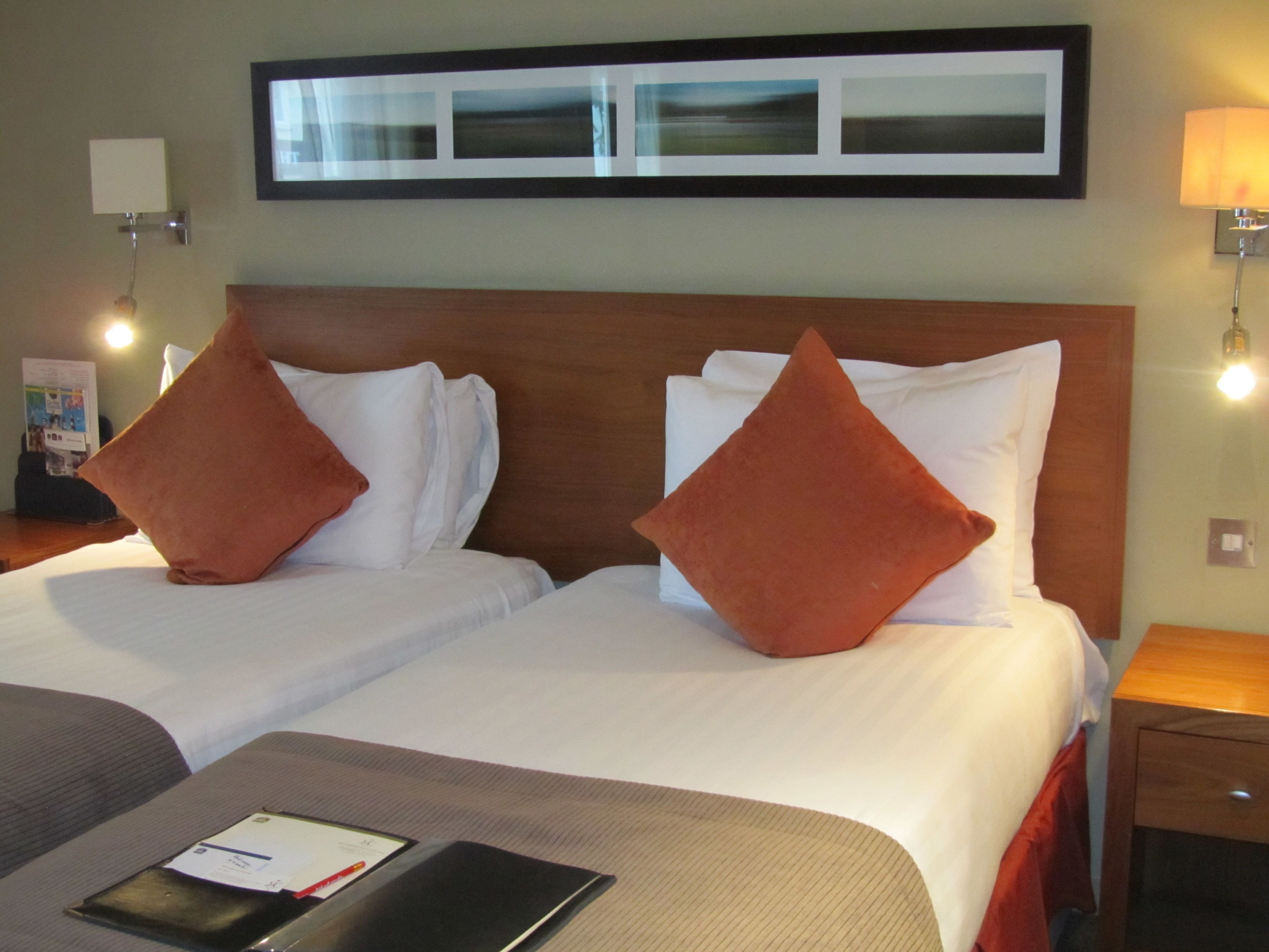 Habitación de hotel con dos camas, cada una con ropa de cama blanca y cabeceros de madera. Hay dos lámparas fijadas a la pared a cada lado de las camas.