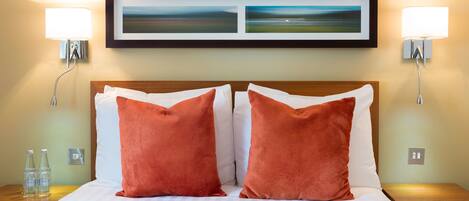 Habitación de hotel con una cama bien hecha, dos almohadas rojas, dos lámparas fijadas a la pared y dos cuadros de paisajes sobre la cama.