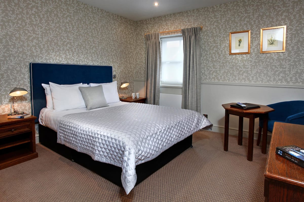 A neatly arranged hotel room with a large bed, a bedside table with a lamp, a small table with a remote control, and a framed picture on the wall.