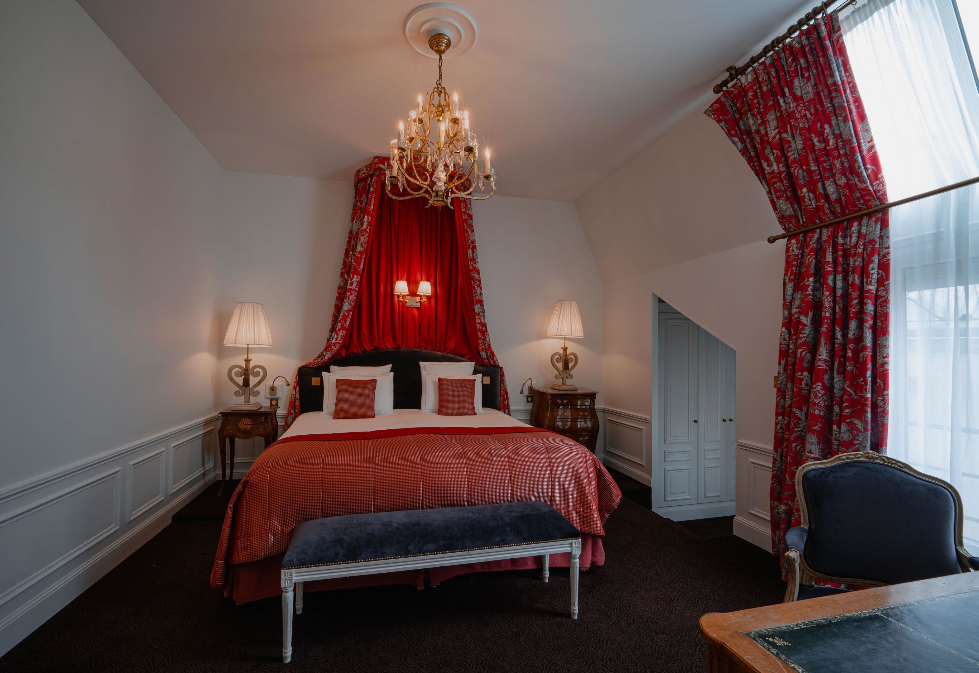 Ein Schlafzimmer mit einem großen Bett, zwei Nachttischen, einer Bank, einem Sessel, einer Deckenleuchte und einem Fenster mit Vorhängen.
