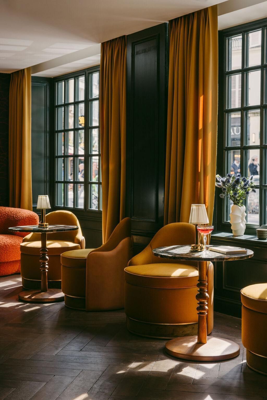 A room with large windows, dark walls, and yellow curtains. There are multiple seating areas with round tables and lamps.