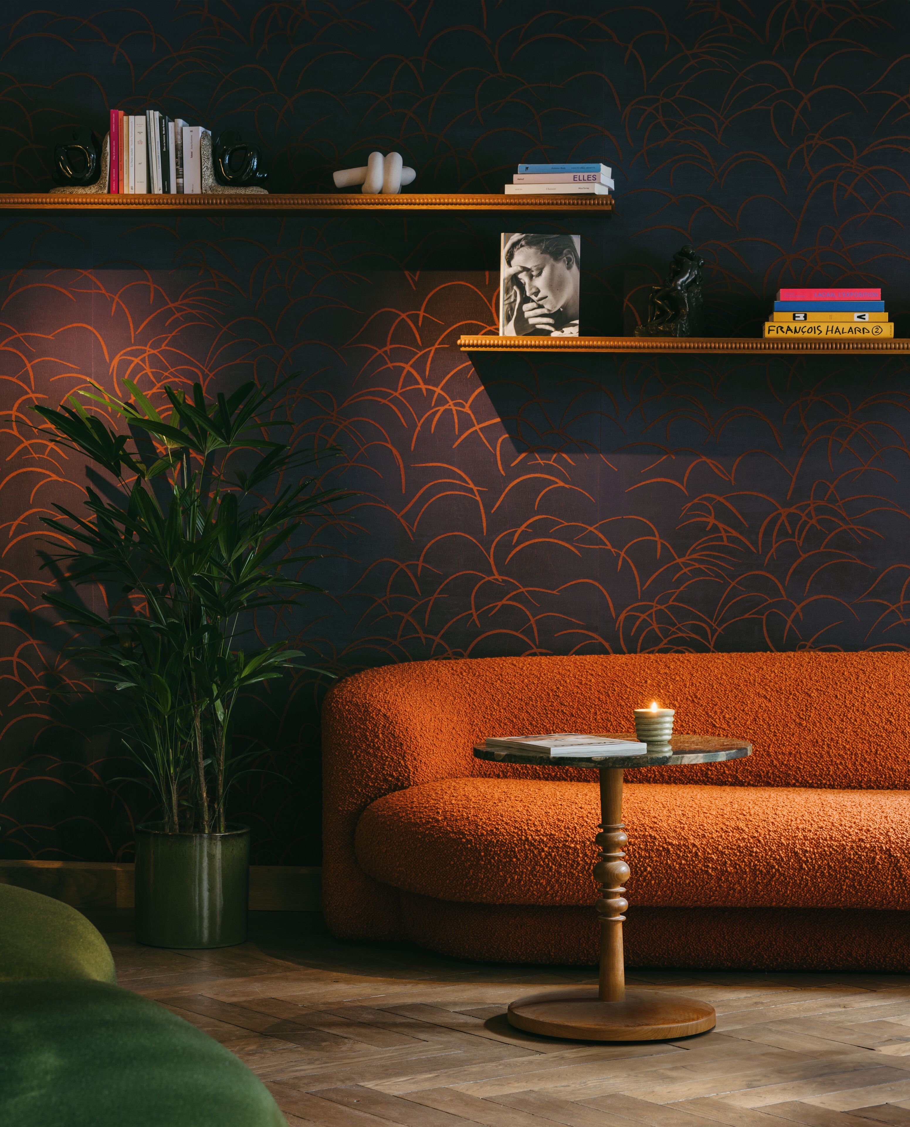 A cozy room with a textured orange sofa, a small round table with a candle, a potted plant, and shelves with books and decorative items.