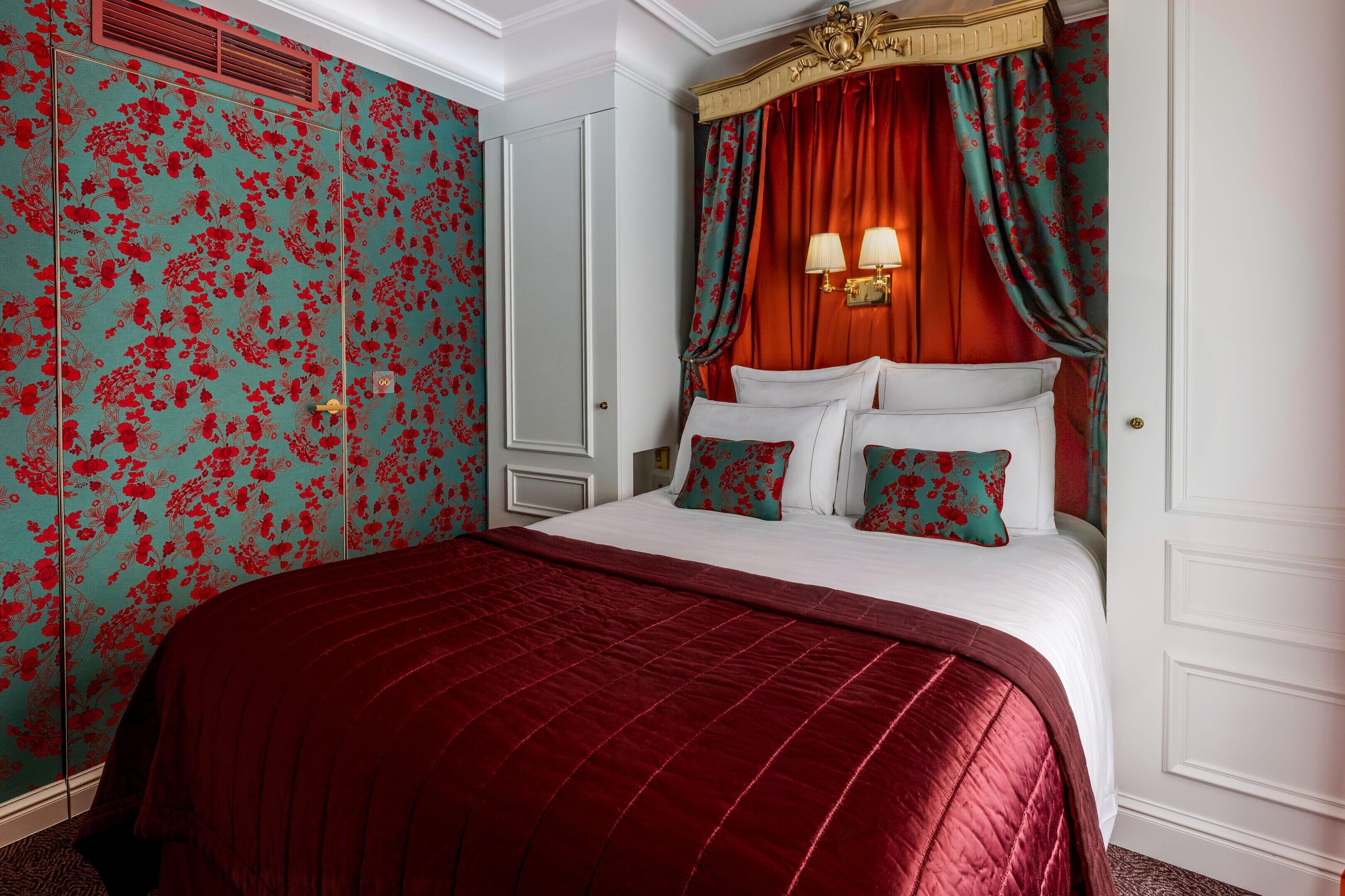 A bedroom with a large bed, red curtains, floral wallpaper, and a white door.