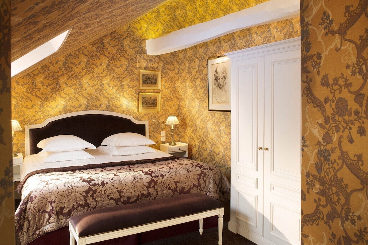 A bedroom with a large bed, a nightstand, a bench, a wardrobe, and framed artwork on the wall.