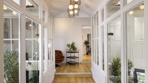 Ein Flur mit Holzboden, weißen Wänden und Glastüren, die in einen Garten führen. Eine schwarze Metallregalgestell präsentiert Pflanzen und ein brauner Stuhl steht daneben.