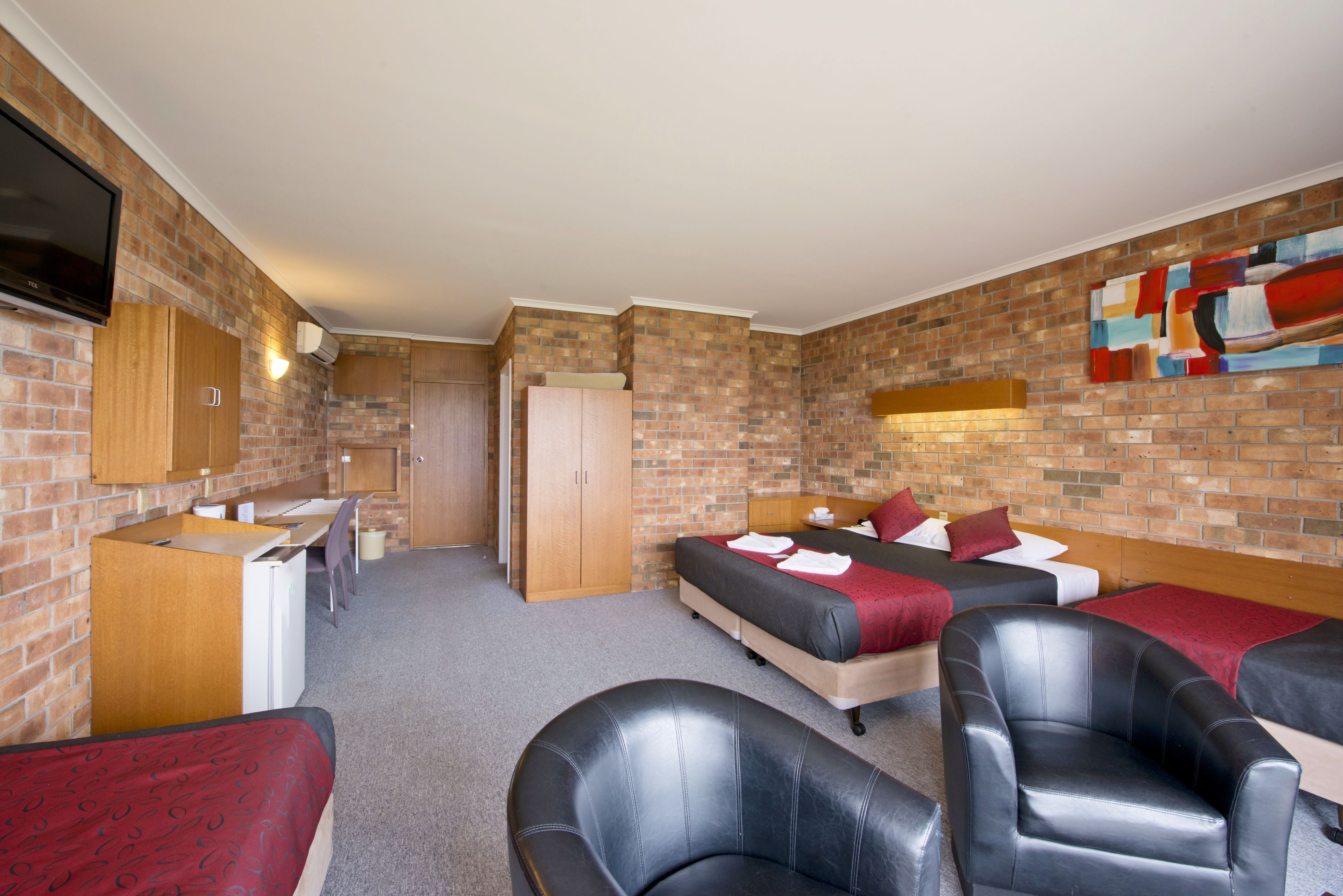 Ein Zimmer mit Backsteinwand, einem Bett mit roter Bettwäsche, einem Schreibtisch mit Computer und einem an der Wand montierten Fernseher.