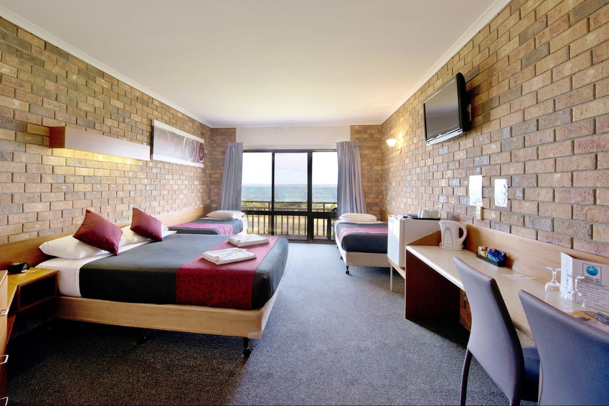 A hotel room with two beds, a desk, a TV, and a balcony with a view of the sea.