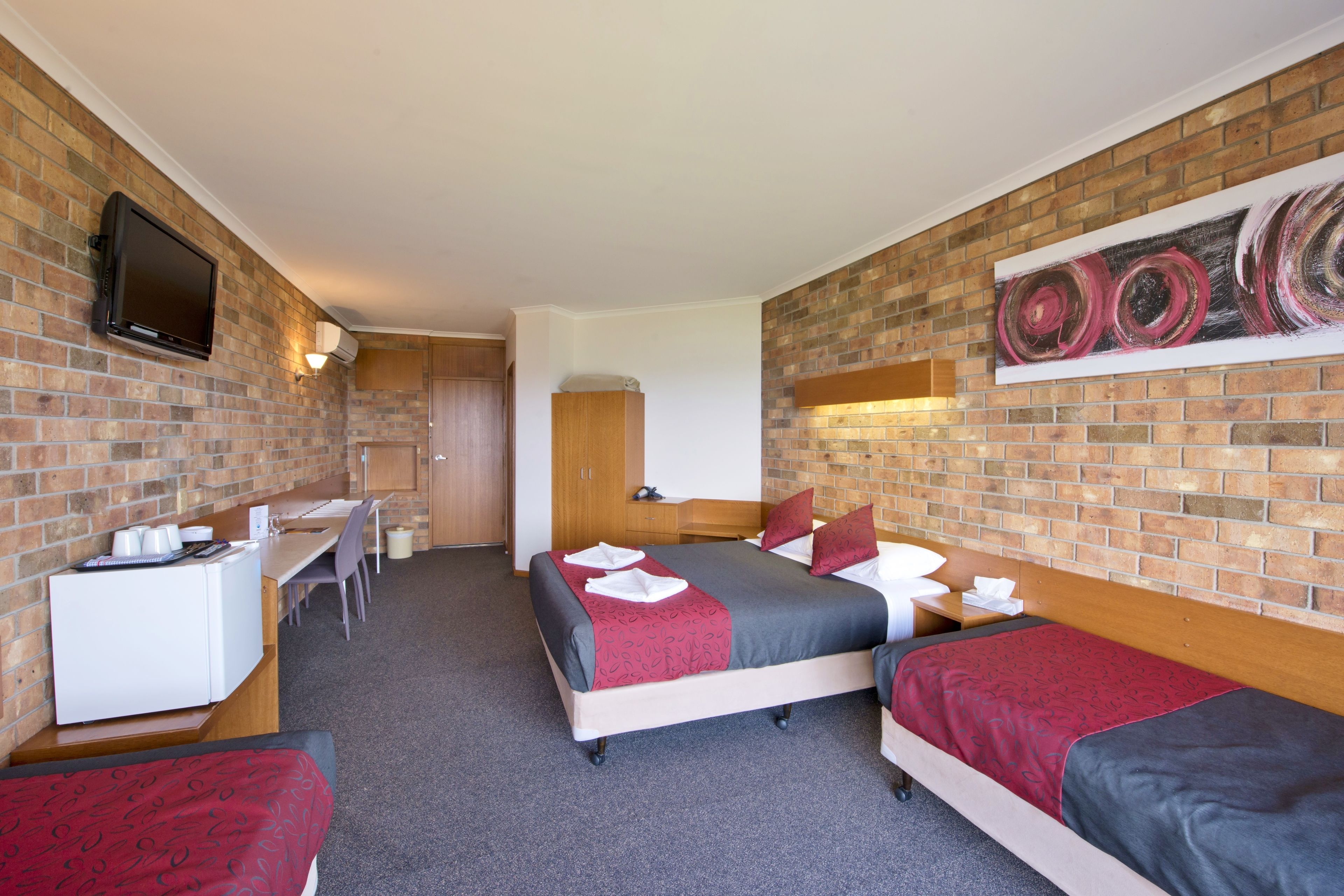 A hotel room with a brick wall, a flat-screen TV mounted above the desk, a bed with a red and grey bedspread, and a painting on the wall.