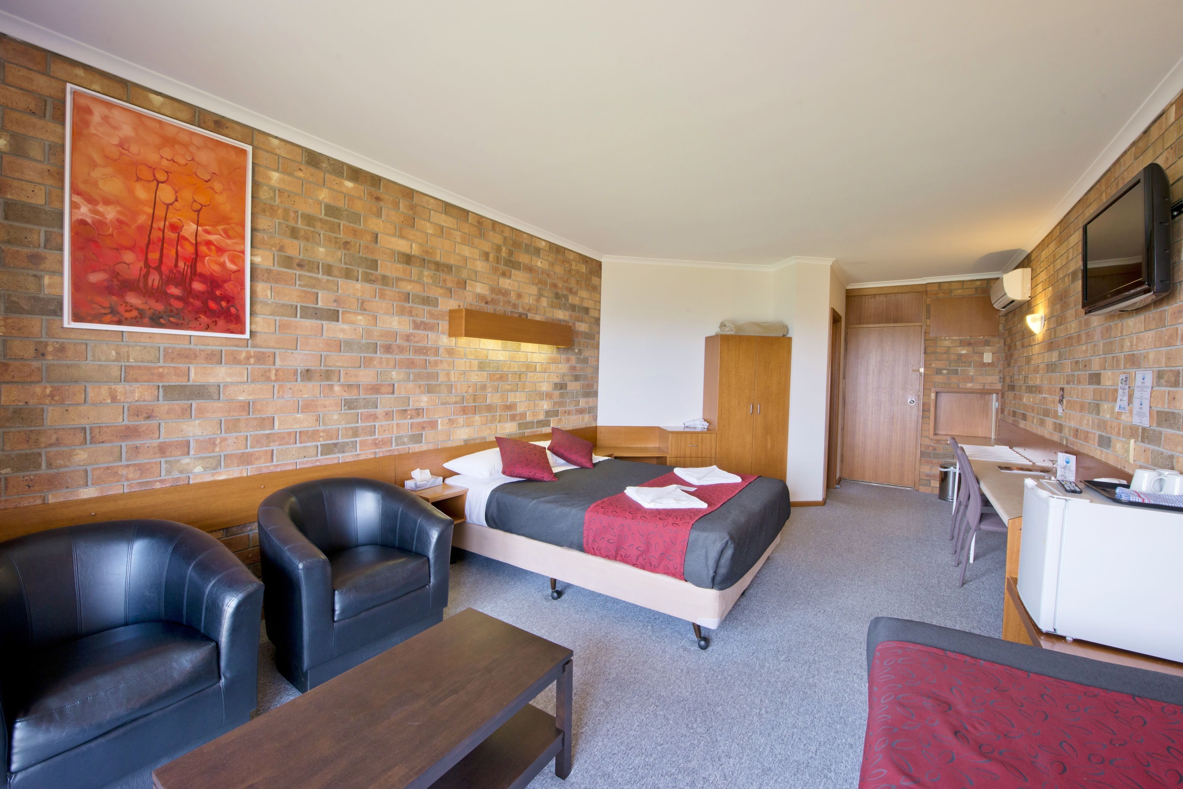 Ein Hotelzimmer mit Backsteinwand, einem Bett mit roter Bettwäsche, einem Schreibtisch, einem Sessel und einem Fernseher.