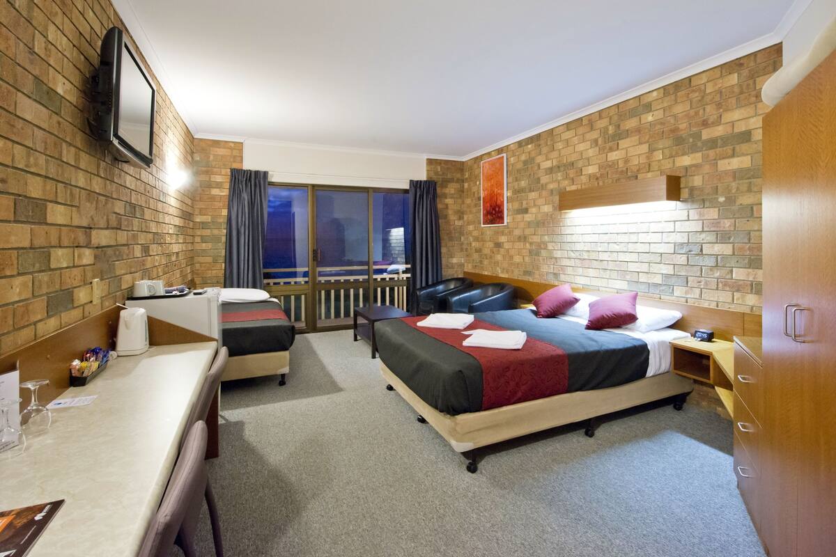 A hotel room with a brick wall, a bed with multiple pillows, a desk with a chair, a small table, and a TV mounted on the wall.