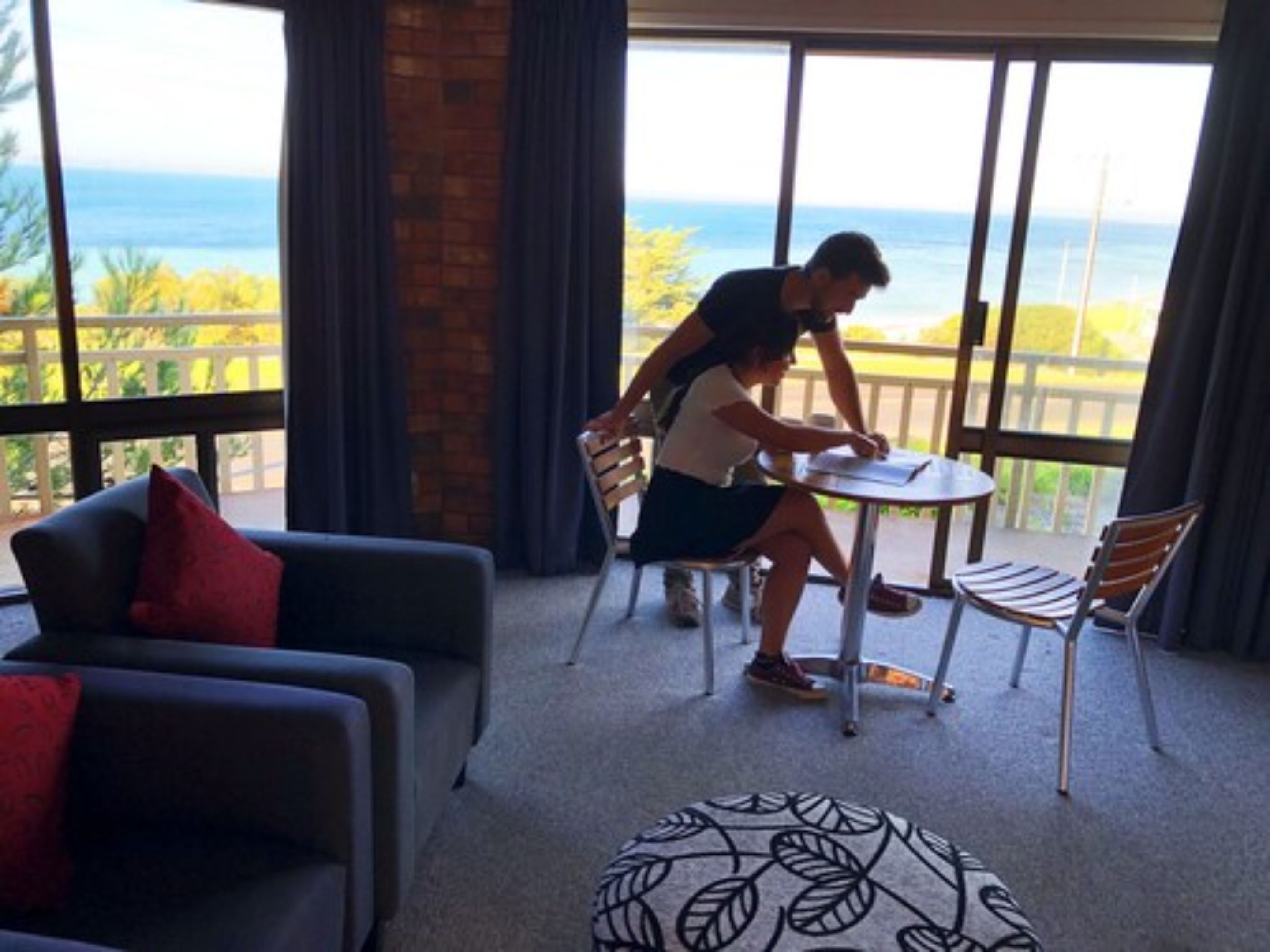 Una habitación con un ventanal grande con vistas a un cuerpo de agua, una mesa redonda y un sofá con un cojín rojo.