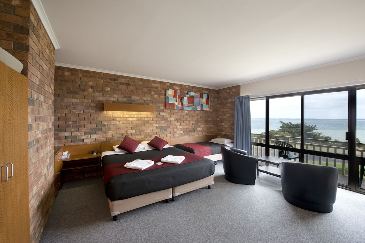 A modern hotel room with a brick wall, two beds, a large window with a view of the sea, and a balcony with a chair.