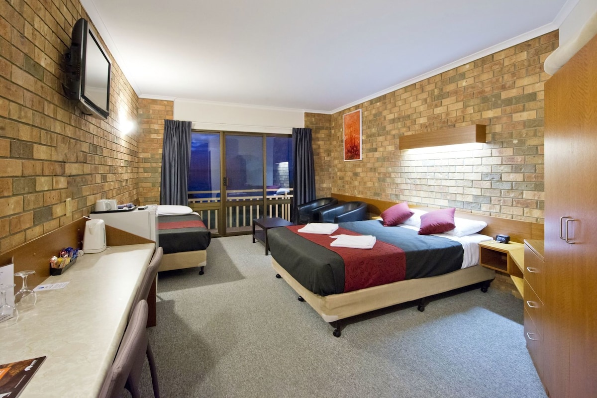 Une chambre d’hôtel avec un mur en briques, un lit recouvert d’une courtepointe rouge et noire, un bureau avec une chaise, une petite table avec une lampe et un téléviseur fixé au mur.