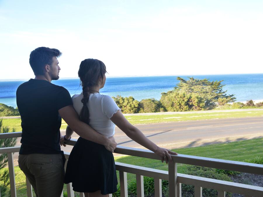 A couple standing on a balcony overlooking a coastal road with a view of the ocean and greenery.