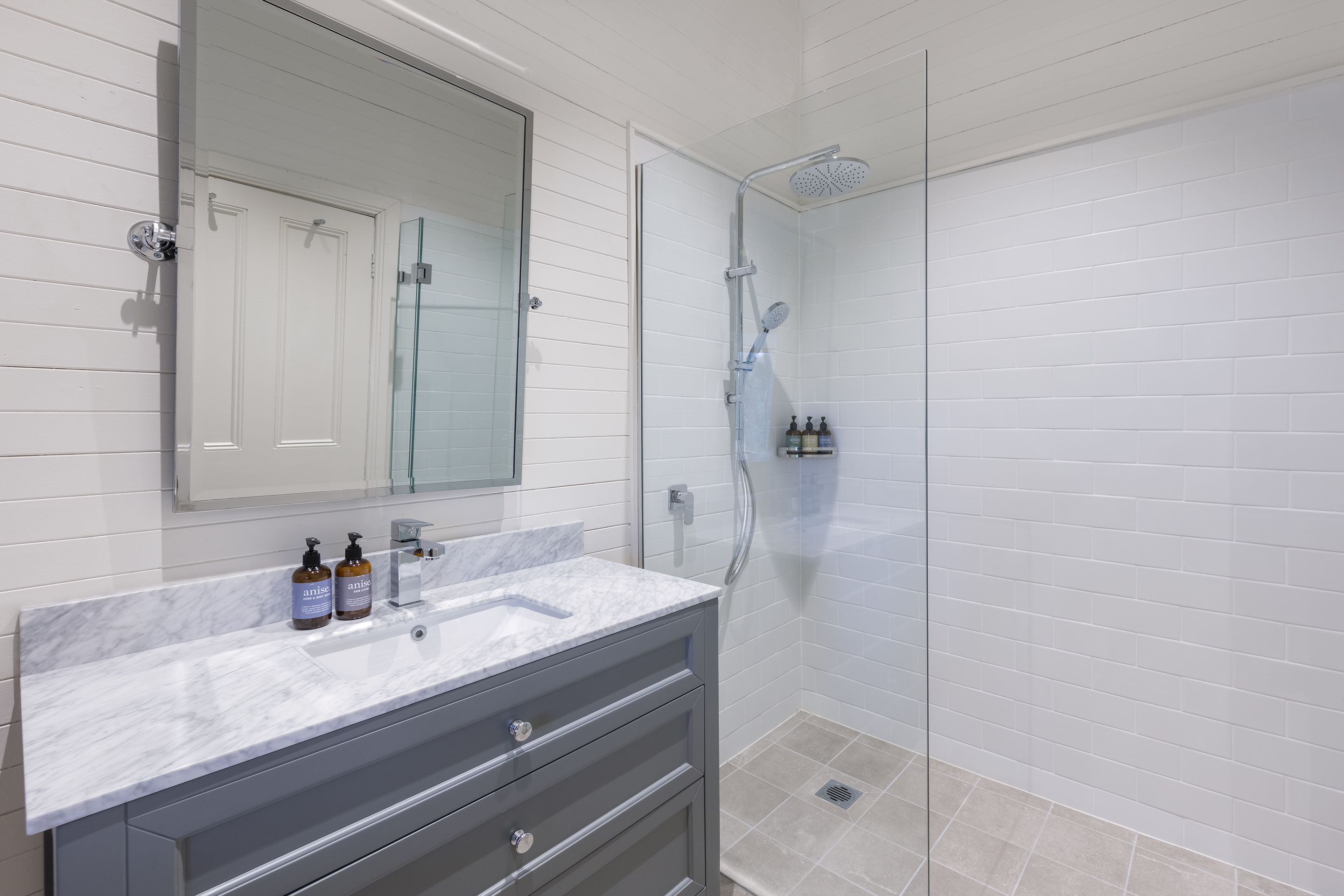 Un bagno con doccia rivestita di piastrelle bianche, box doccia in vetro, lavabo in marmo e uno specchio sopra.