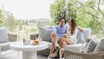 A couple sitting on a wicker sofa with a table between them, a white tablecloth, and a view of greenery in the background.