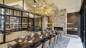 A modern dining area with a long wooden table set for a meal, a stone fireplace, and a view of a wine cellar. - Spicers Guesthouse (Pokolbin)