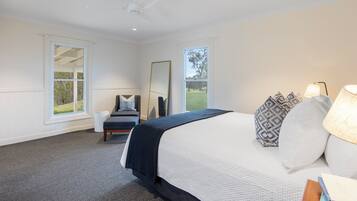 A bedroom with a large bed, a bedside table with a lamp, a chair, a mirror, and a window with a view of greenery.