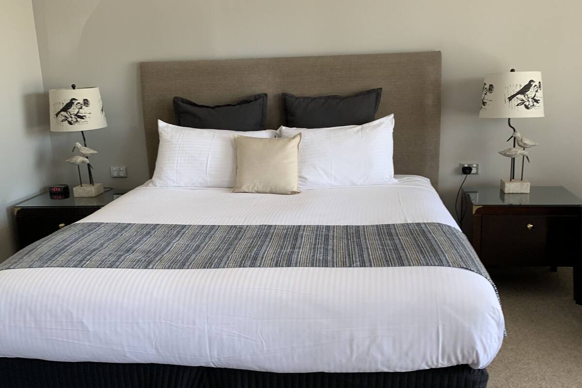 A neatly made bed with a grey and white striped bedspread, two pillows, and a dark skirted end table with a lamp.