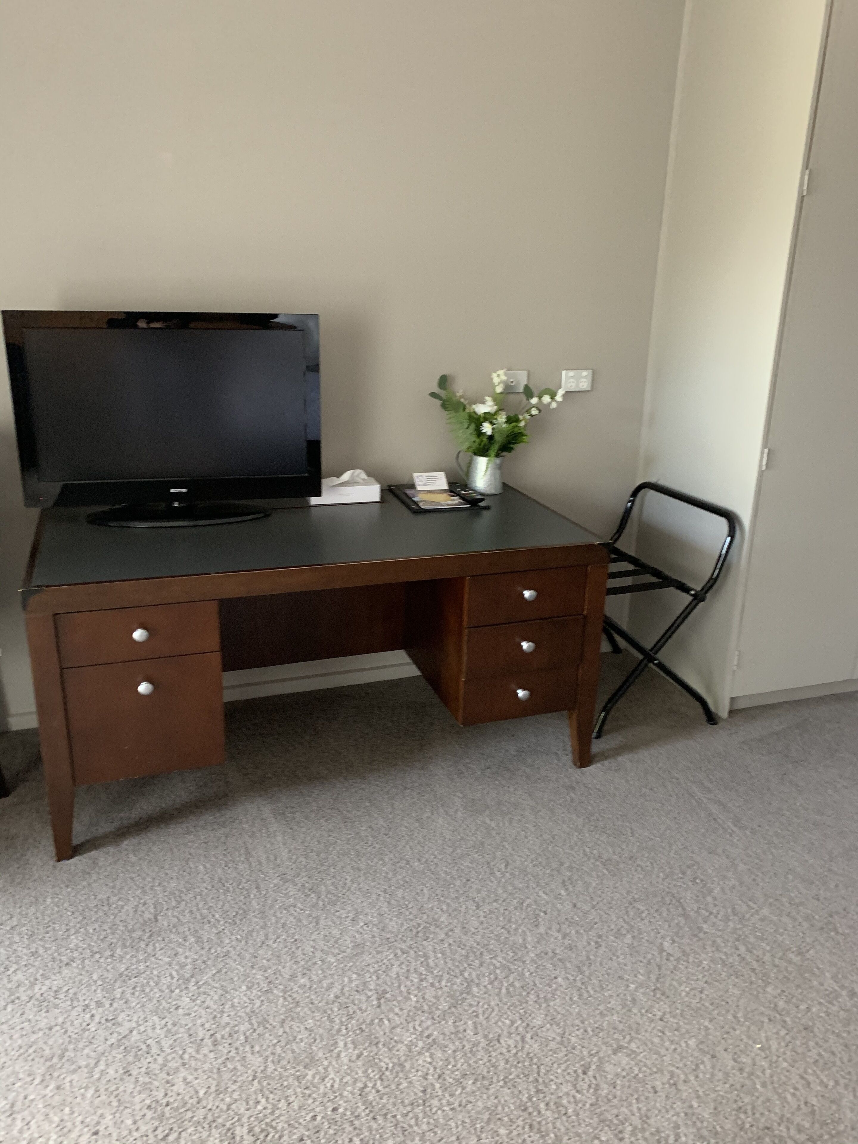 A wooden desk with a flat-screen TV, a vase with flowers, and a chair.
