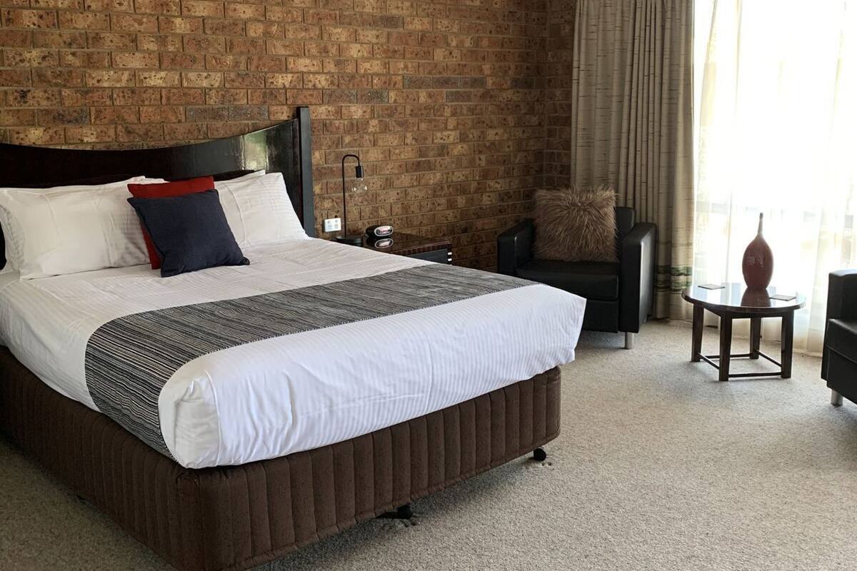 A hotel room with a brick wall, a large bed with white bedding, a nightstand, a chair, and a small table.