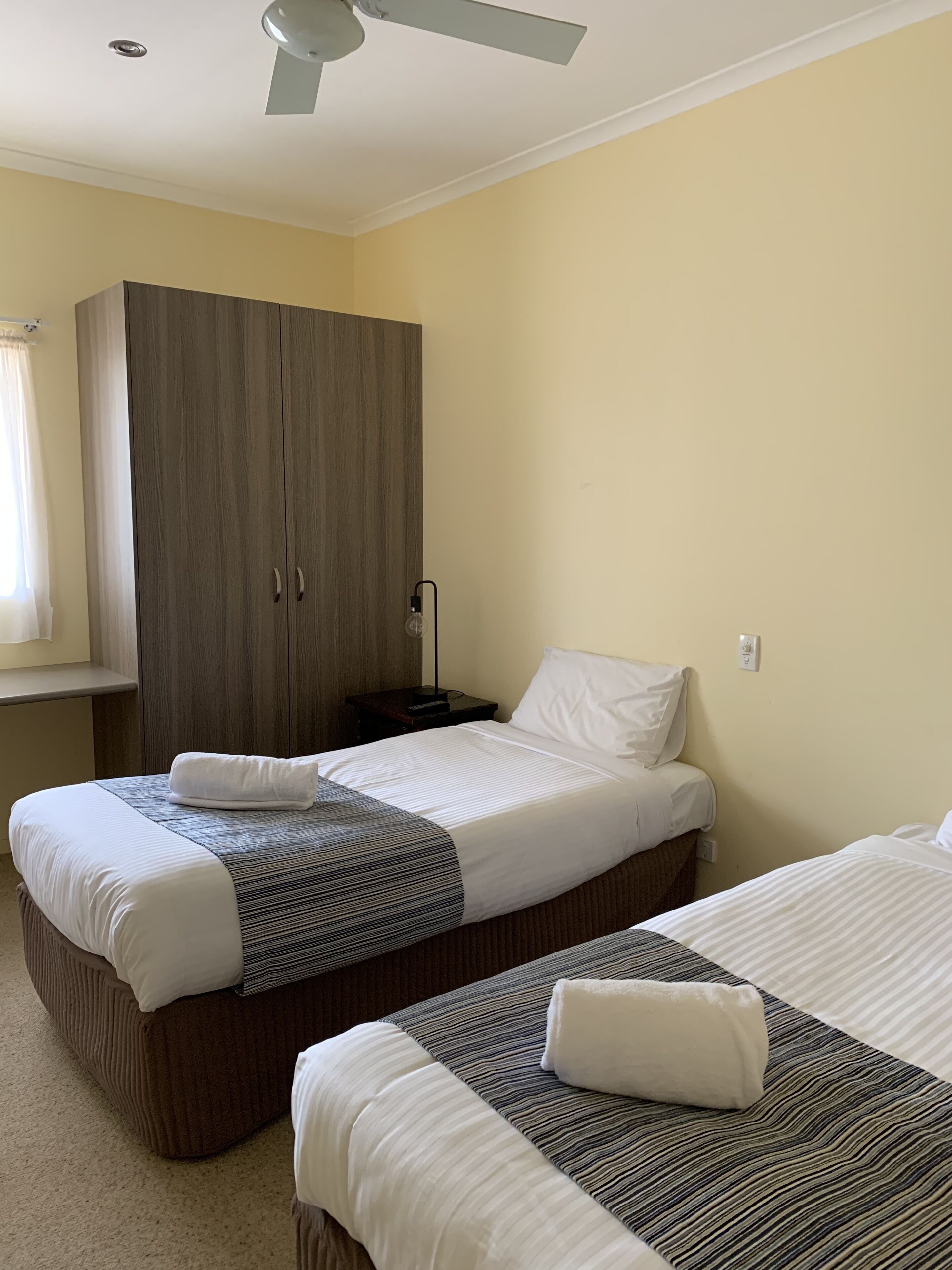 A hotel room with two beds, a wardrobe, a ceiling fan, and a bedside table with a lamp.