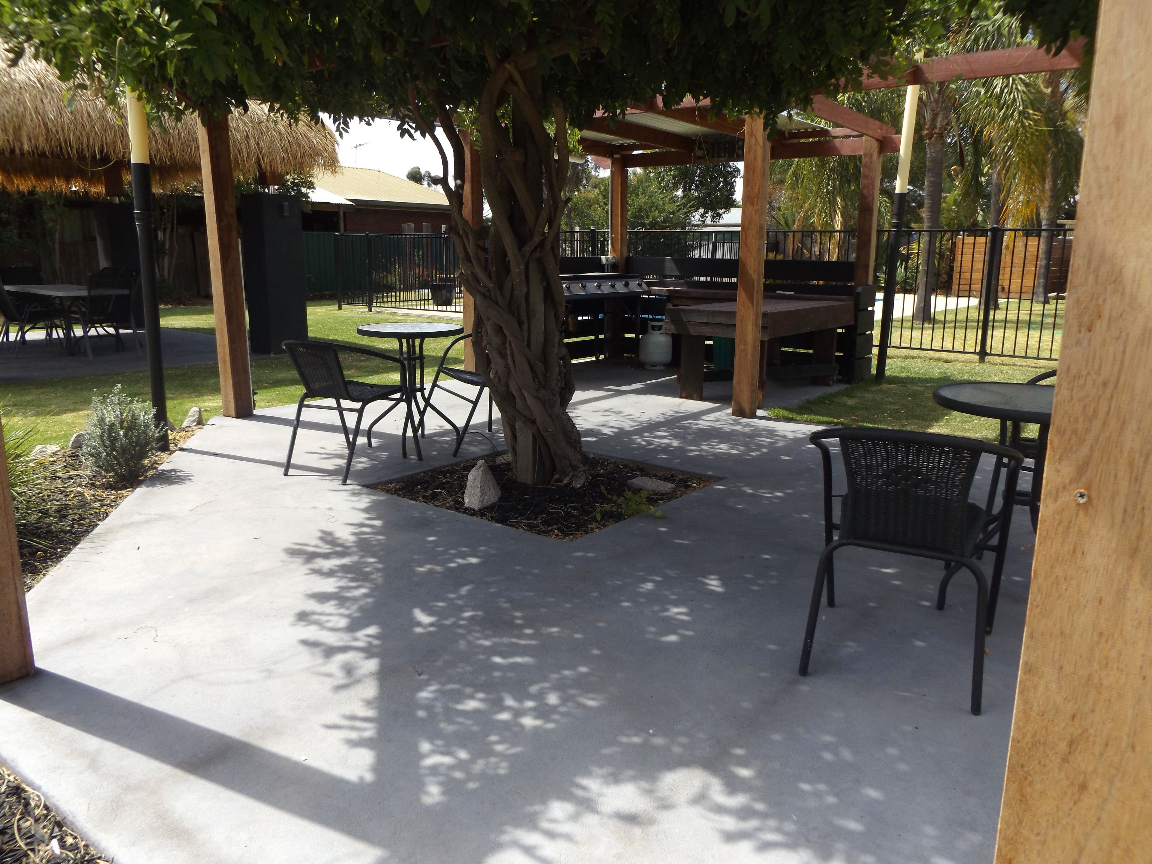 Un patio al aire libre con mesas y sillas bajo una pérgola, rodeado de una valla y vegetación.