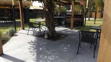 Un patio al aire libre con mesas y sillas bajo una pérgola, rodeado de una valla y vegetación.