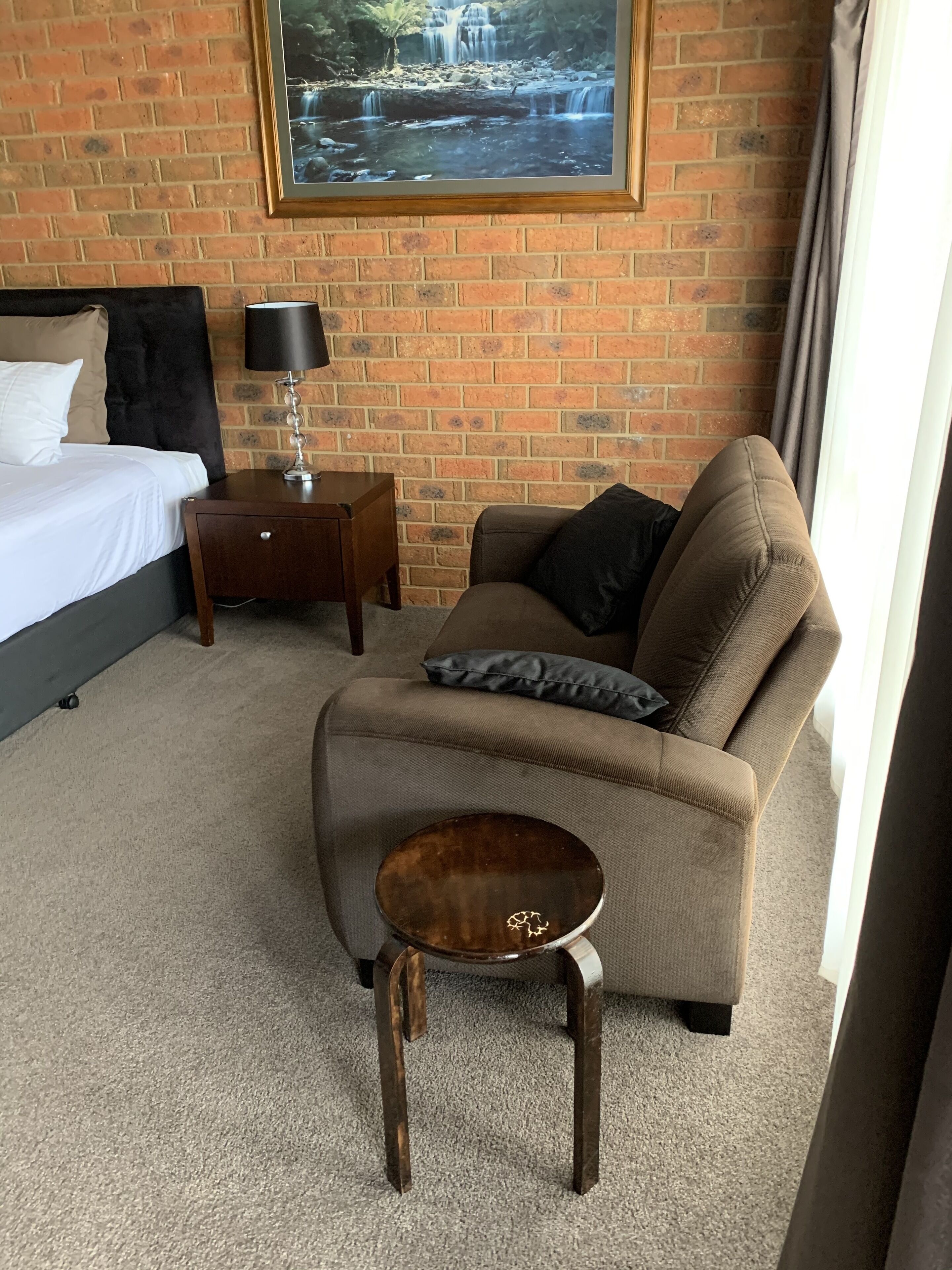 A hotel room with a brick wall, a bed with white linens, a brown sofa with a pillow, a small wooden side table, and a framed picture of a waterfall.