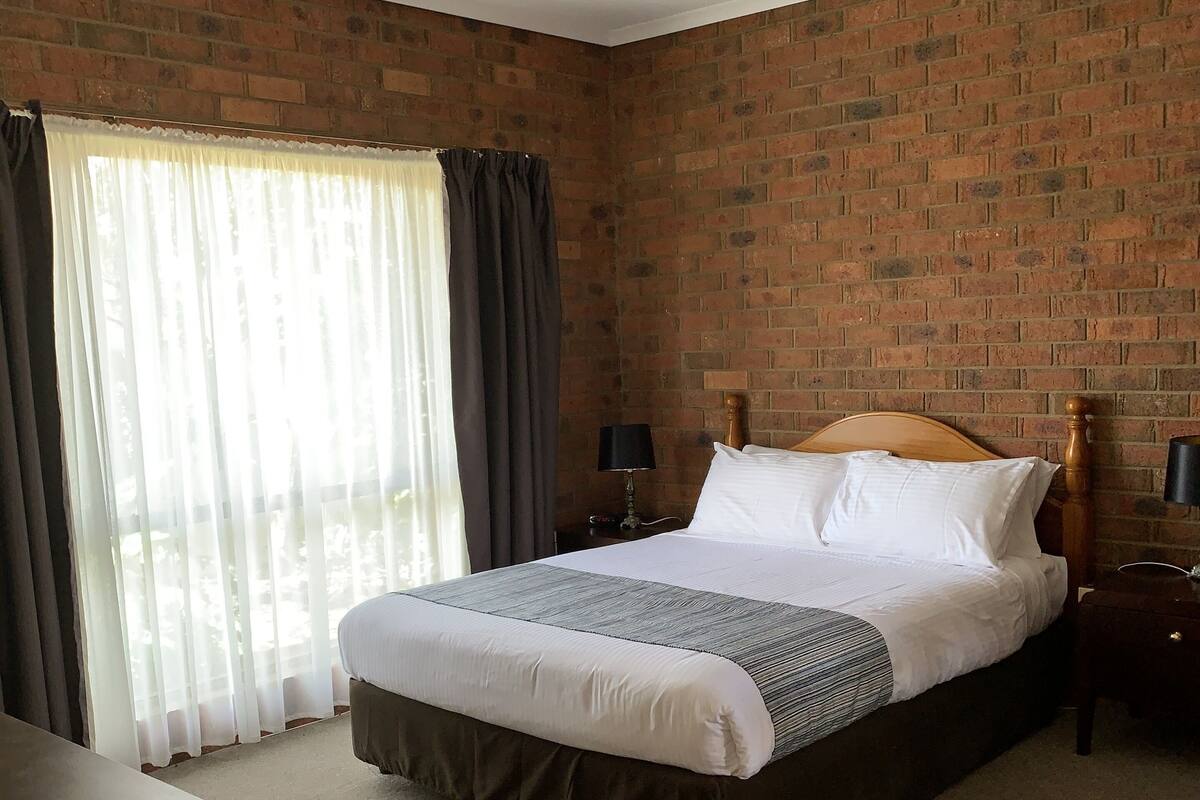 A hotel room with a brick wall, a large bed with white bedding, a wooden headboard, a nightstand with a lamp, and a view through sheer curtains.