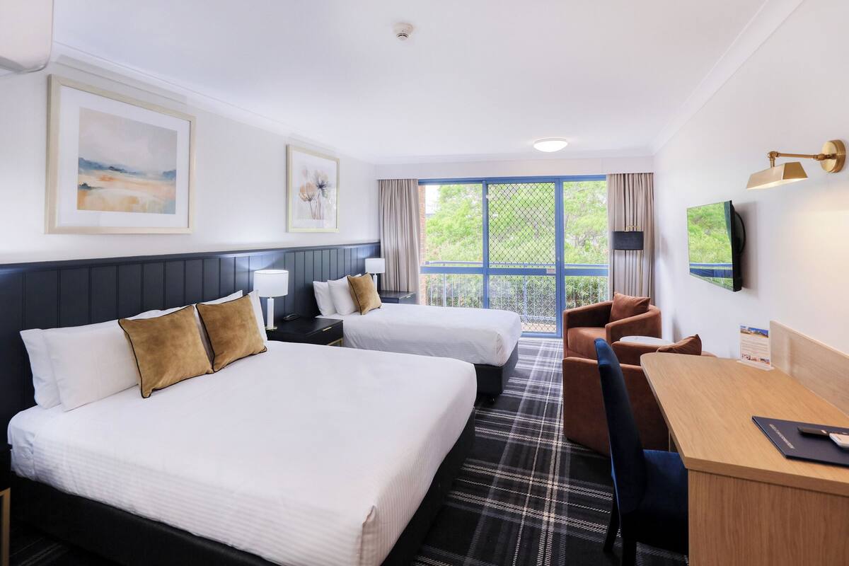 Une chambre d’hôtel avec deux lits, un bureau, une chaise, une télévision et une fenêtre donnant sur un espace verdoyant.