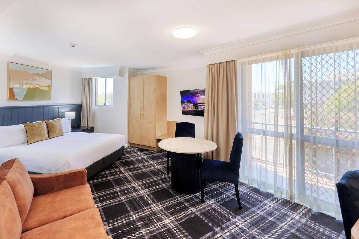 Une chambre d’hôtel équipée d’un lit, d’un bureau, d’une chaise, d’une télévision et d’une grande fenêtre donnant sur un paysage agréable.